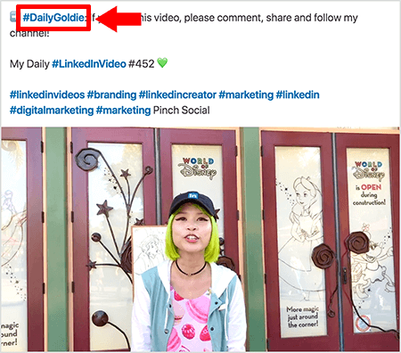 Questo è uno screenshot che illustra come Goldie Chan utilizza gli hashtag nel testo dei suoi post video su LinkedIn. I callout rossi indicano l'hashtag #DailyGoldie nel testo, che è unico per i suoi post video e la aiuta a tenere traccia delle condivisioni. Il post include anche altri hashtag rilevanti che aiutano le persone a trovare il suo video, incluso #LinkedInVideo. Nell'immagine video, Goldie si trova davanti ad alcune porte in uno spettacolo di World of Disney. È una donna asiatica con i capelli verdi. Indossa un berretto LinkedIn nero, una collana girocollo nera, una maglietta rosa con stampa macaron e una giacca blu e bianca.