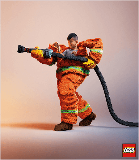 Questa è una foto di una pubblicità LEGO che mostra un giovane ragazzo asiatico all'interno di un'uniforme da pompiere fatta di LEGO. L'uniforme è arancione con una striscia verde neon intorno ai polsini del cappotto e dei pantaloni. Il vigile del fuoco è in piedi con un piede indietro e tiene in mano una manichetta antincendio, anch'essa di lego. La testa del ragazzo spunta dalla parte superiore dell'uniforme, che è molto più grande di lui e si ferma intorno alle spalle. La foto è stata scattata su un semplice sfondo neutro. Il logo LEGO appare in un riquadro rosso in basso a destra. Talia Wolf afferma che LEGO è un ottimo esempio di un marchio che utilizza le emozioni nella pubblicità.
