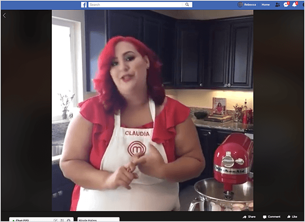 Questo è uno screenshot di un video di Facebook Live con la chef Claudia Sandoval, che ha ospitato nel 2016 come parte di una promozione incrociata con l'app T-Mobile Tuesdays. Nel video, Claudia è in una cucina con armadi neri e ripiani in granito. Una finestra sopra il lavandino lascia entrare la luce naturale nella stanza. Claudia è in piedi accanto a un robot da cucina rosso. È una donna latina con i capelli rosso vivo che le cadono appena sotto le spalle. È truccata, un top rosso acceso e un grembiule bianco con Claudia e il logo MasterChef ricamato in filo rosso. Mentre parla, fa dei gesti con le mani. Nel 2016, Stephanie Liu ha collaborato con Claudia per produrre questo video dal vivo.